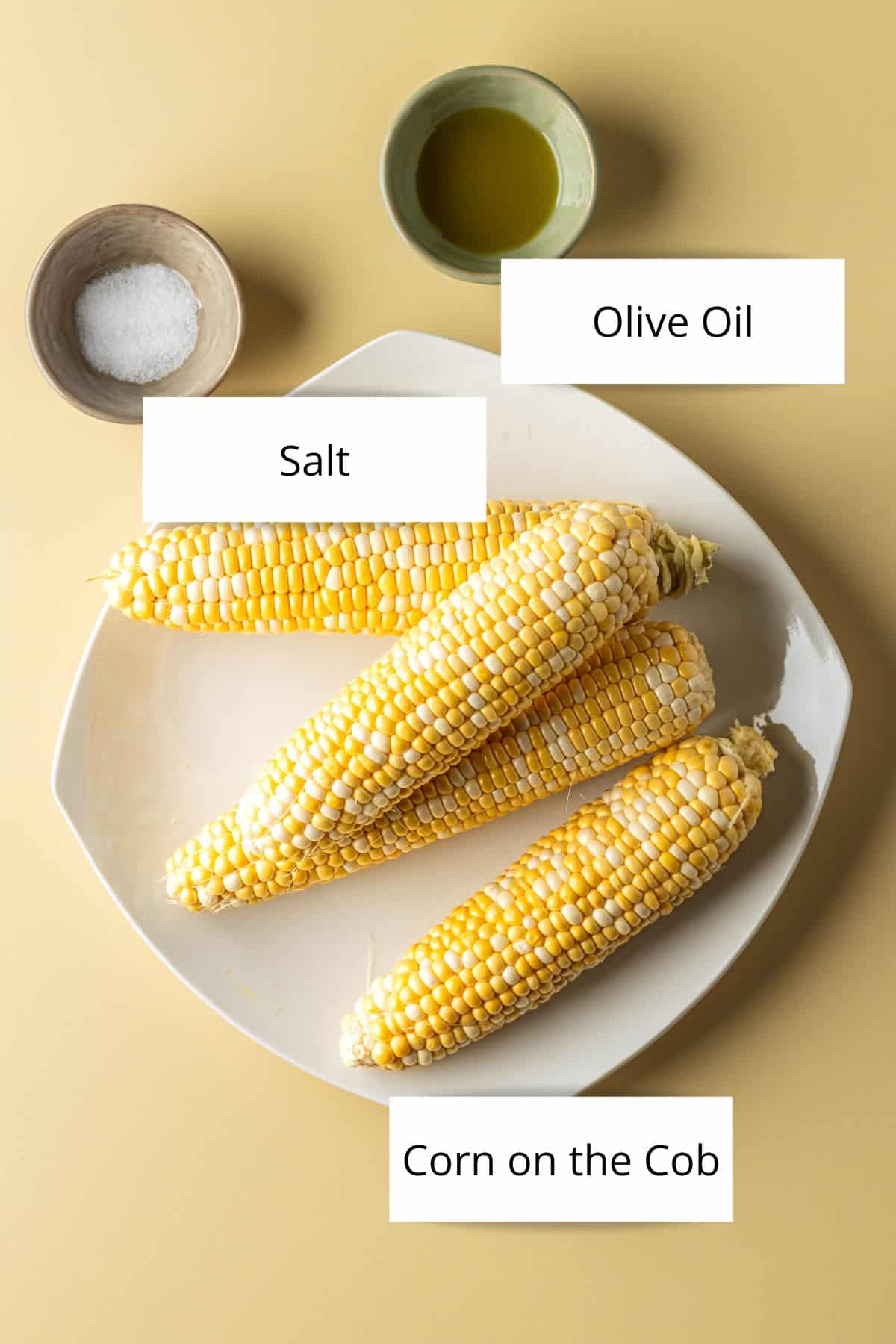 Four ears of corn on a square white plate with a small bow of salt and a small bowl of olive oil.