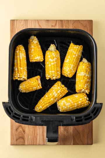 Cooked ears of corn in an air fryer basket on a wooden cutting board.