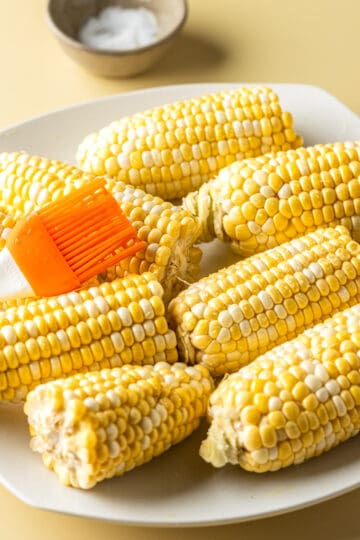 Corn on the cob on a white plate being brushed with oil from a silicone orange brush.