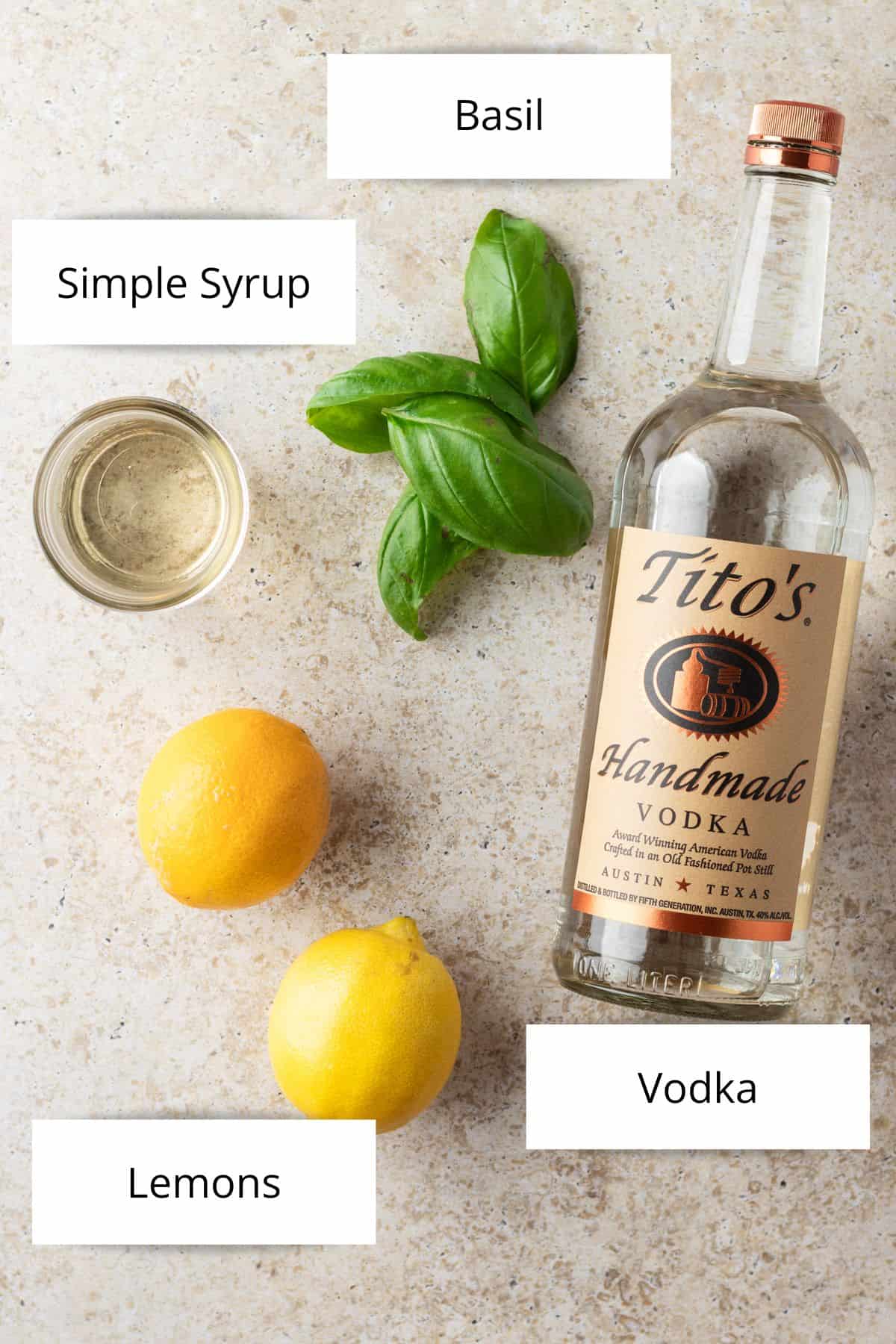 A bottle of Tito's vodka, two lemons, a jar of simple syrup and four basil leaves.