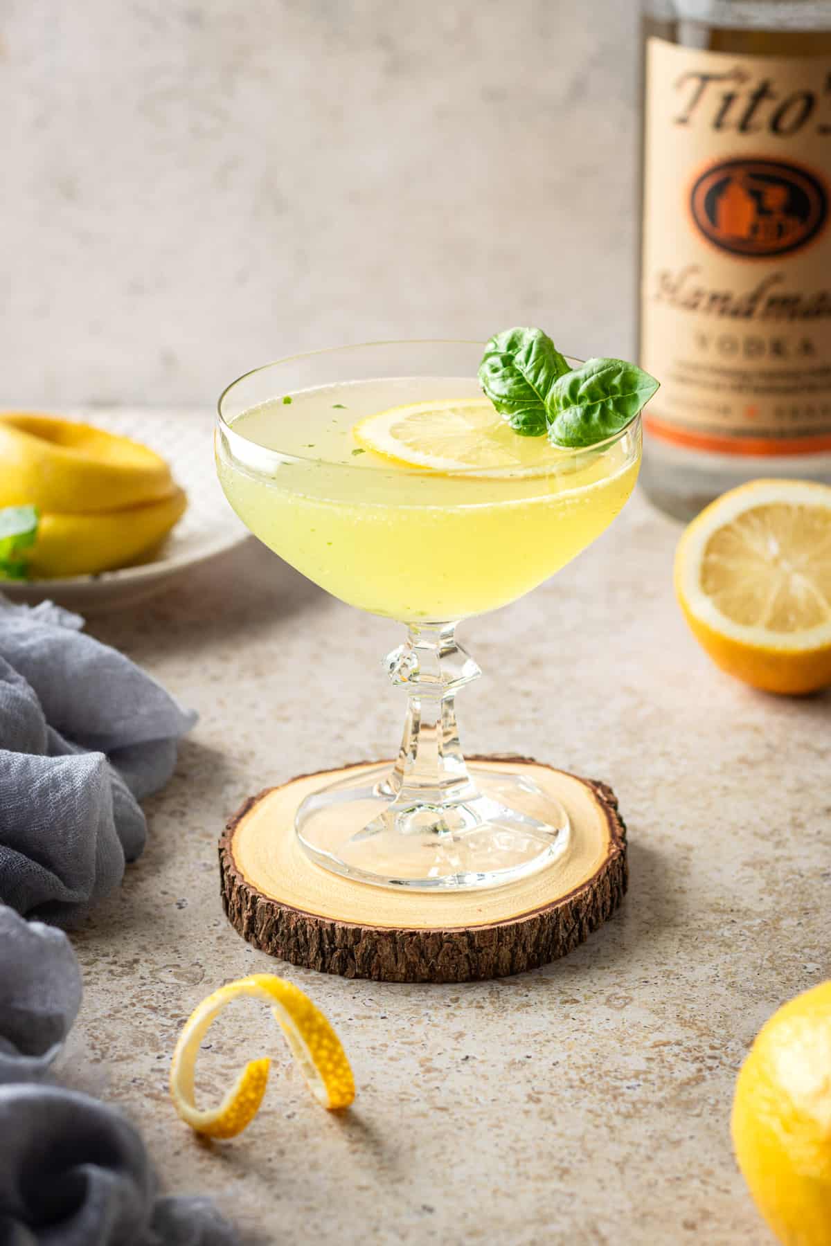 A lemon drop basil martini on a wooden coaster with a bottle of Tito's vodka in the background garnished with basil and lemons.