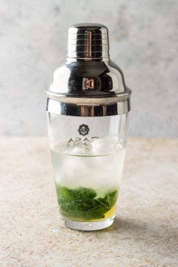 A clear cocktail shaker with fresh basil, lemon juice, ice, vodka and simple syrup.