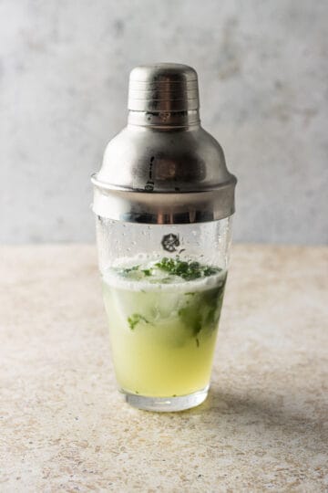 A clear cocktail shaker after being shaken with muddle basil, lemon juice, vodka and ice.