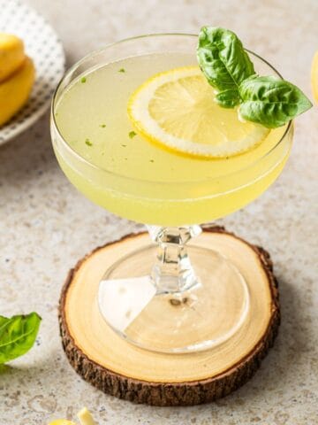 A lemon basil vodka martini in a coupe glass on a wooden coaster garnished with lemon and basil.