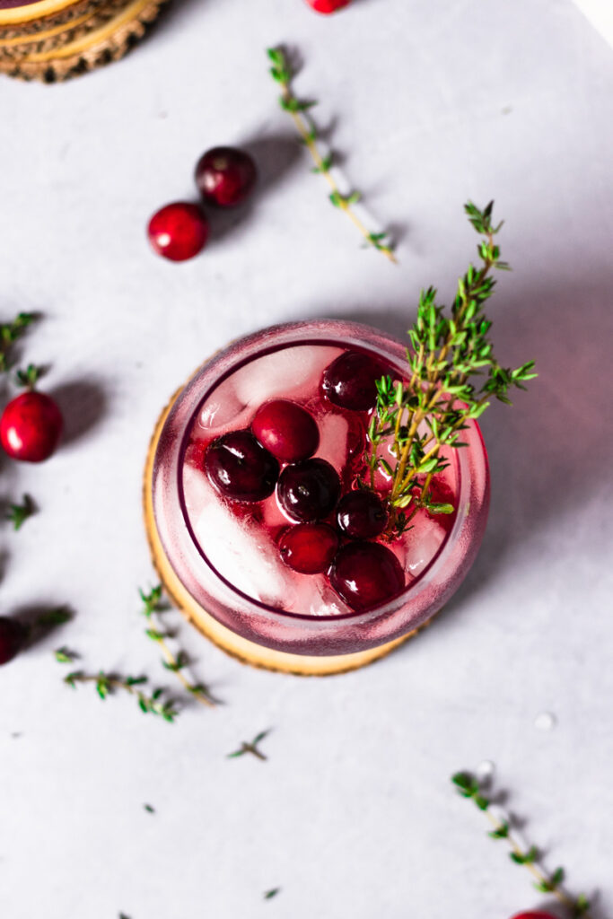 Overhead of a wine glass with a cranberry gin cocktail garnished with fresh cranberries and a thyme sprig. 