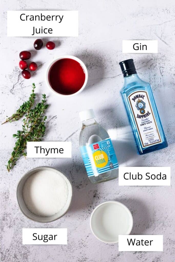 Overhead of a small bowl of cranberry juice, a bottle of gin, thyme sprigs, cane sugar, water and club soda. 