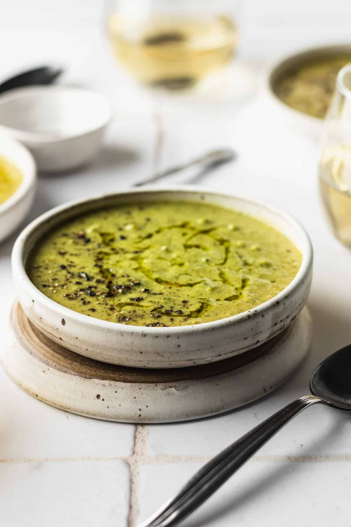 A white bowl of broccoli zucchini soup with a spoon and white wine.