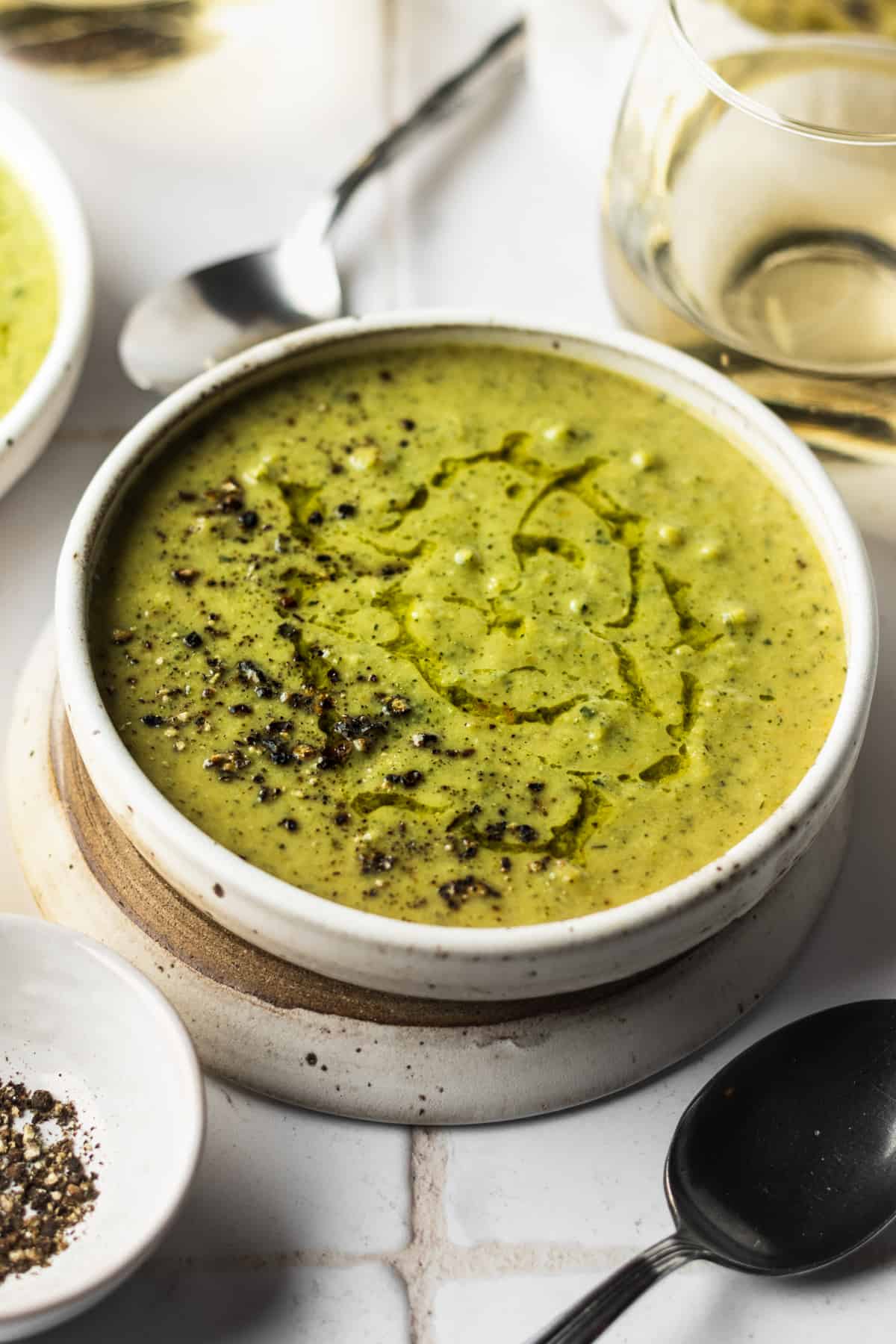 A white bowl of broccoli zucchini soup with a spoon, white wine and black pepper.