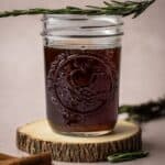 A mason jar with brown sugar spiced simple syrup on a rustic wooden coaster with a sprig of rosemary.