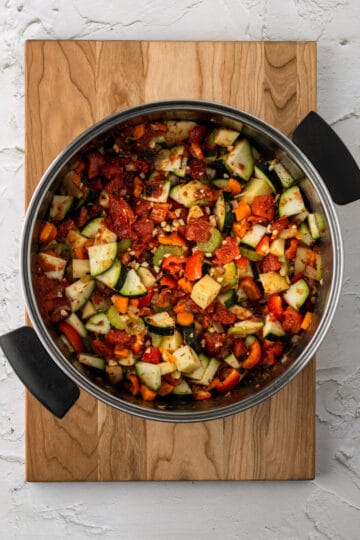 Large pot on a cutting board withs sautéed onion, carrots, celery, garlic, potatoes, zucchini and fire roasted tomatoes.