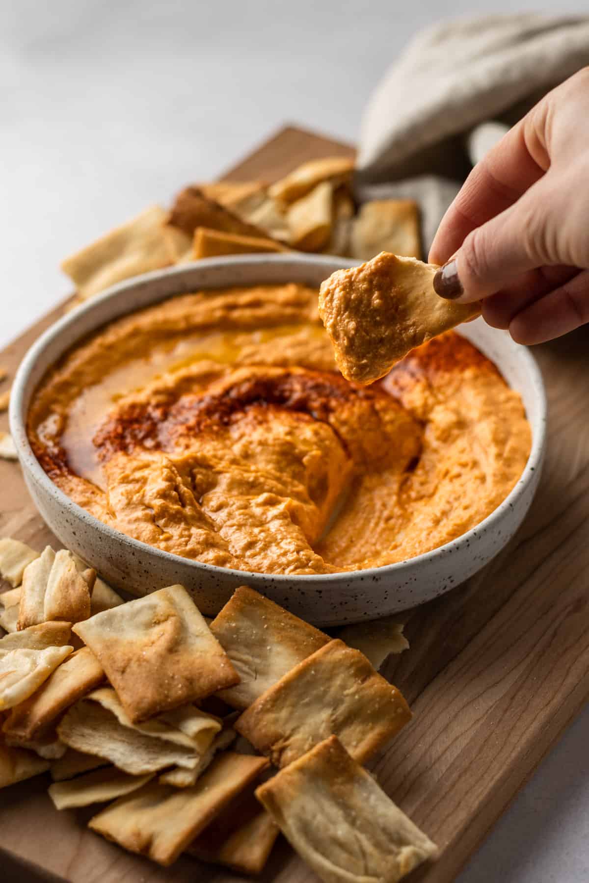 White bean buffalo hummus in a white bowl on a wooden cutting board with a hand scooping out hummus on a pita chip.