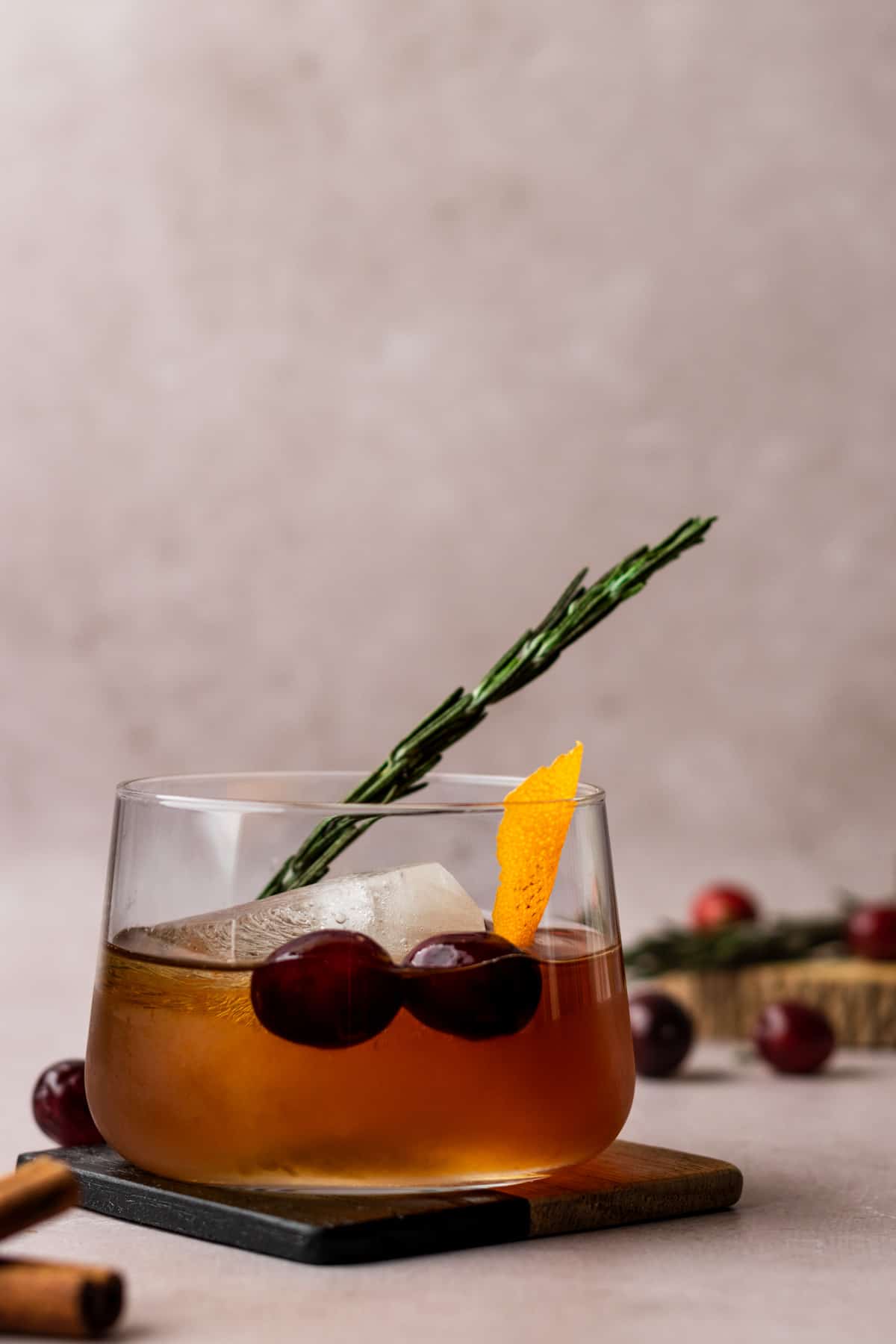 Winter old fashioned cocktail on a square coaster garnished with cranberries, rosemary and orange peel.