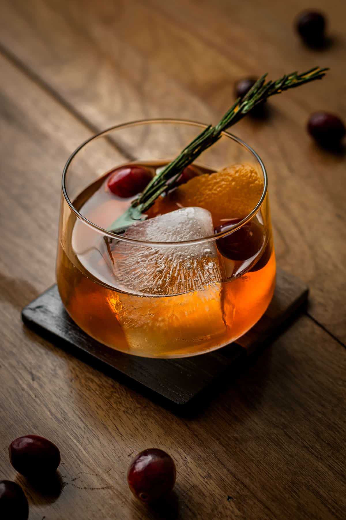 Winter old fashioned cocktail on a square coaster garnished with cranberries, rosemary and orange peel.