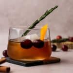 Winter old fashioned cocktail on a square coaster garnished with cranberries, rosemary and orange peel.