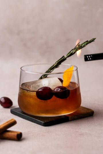 A winter old fashioned in a whiskey glass on a coaster with a rosemary sprig being lit on fire.