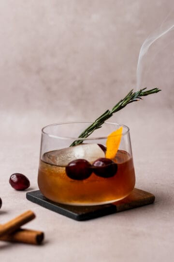 A winter old fashioned in a whiskey glass on a coaster with a smoking rosemary sprig.