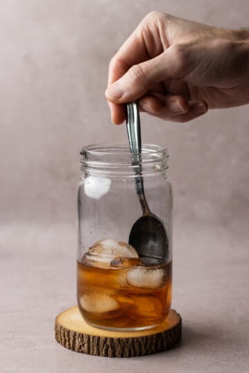 A jar on a wooden coaster with ice, bourbon, simple syrup and bitters being stirred with a spoon.