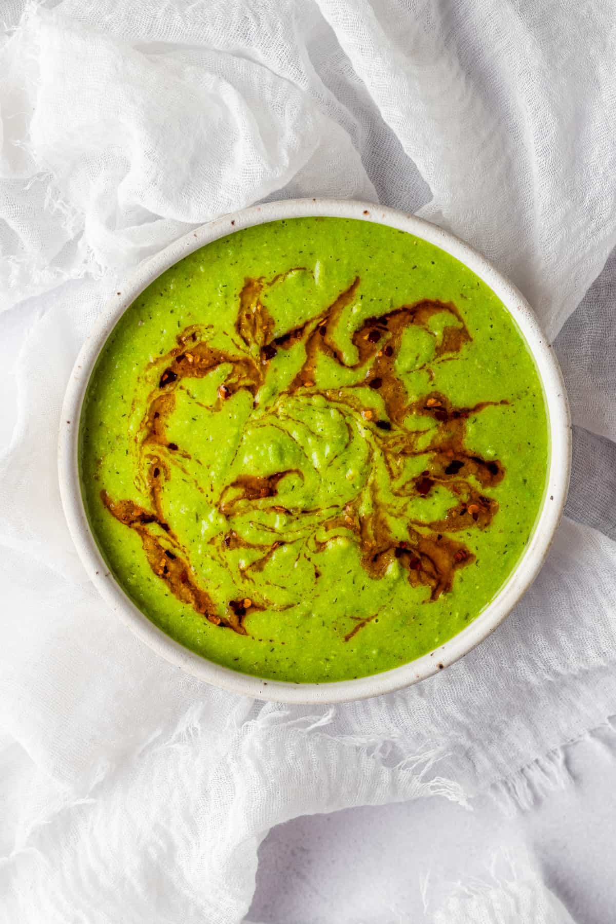 A white bowl on a linen with creamy green pea soup and garlic chili oil drizzled on top.