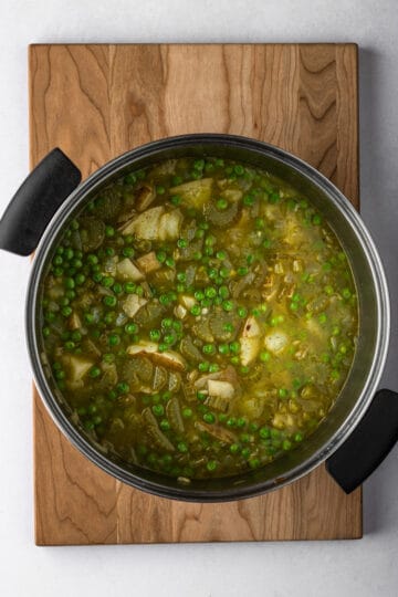 A large pot with sautéed vegetables, peas potatoes, spices and broth on a wooden cutting board.