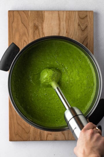 A large pot of green pea soup on a wooden cutting board being blended with a hand held immersion blender.