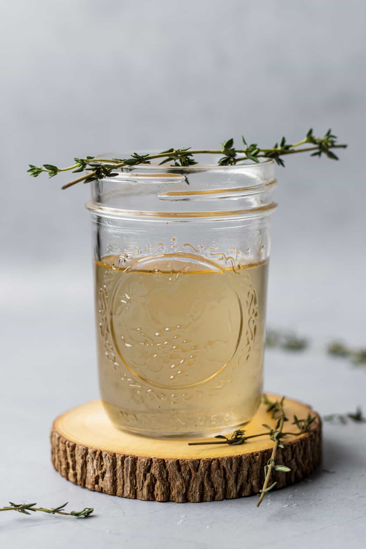A small mason jar on a wooden coaster filled with thyme simple syrup and garnished with thyme leaves.