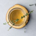 Overhead view of a mason jar on a wooden coaster filled with thyme simple syrup and garnished with thyme leaves.