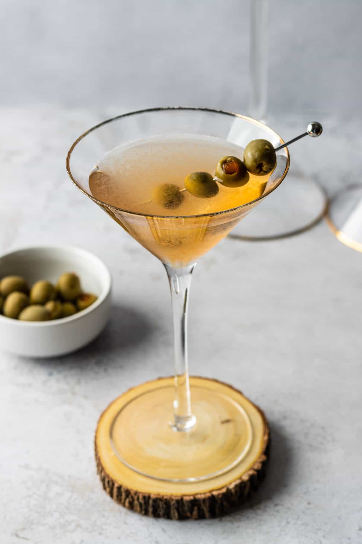 A spicy dirty martini garnished with four green olives on a wooden coaster with a small white bowl of extra green olives.