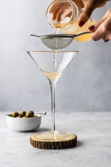 Hot and dirty martini being poured into a martini glass and strained using a fine mesh strainer.