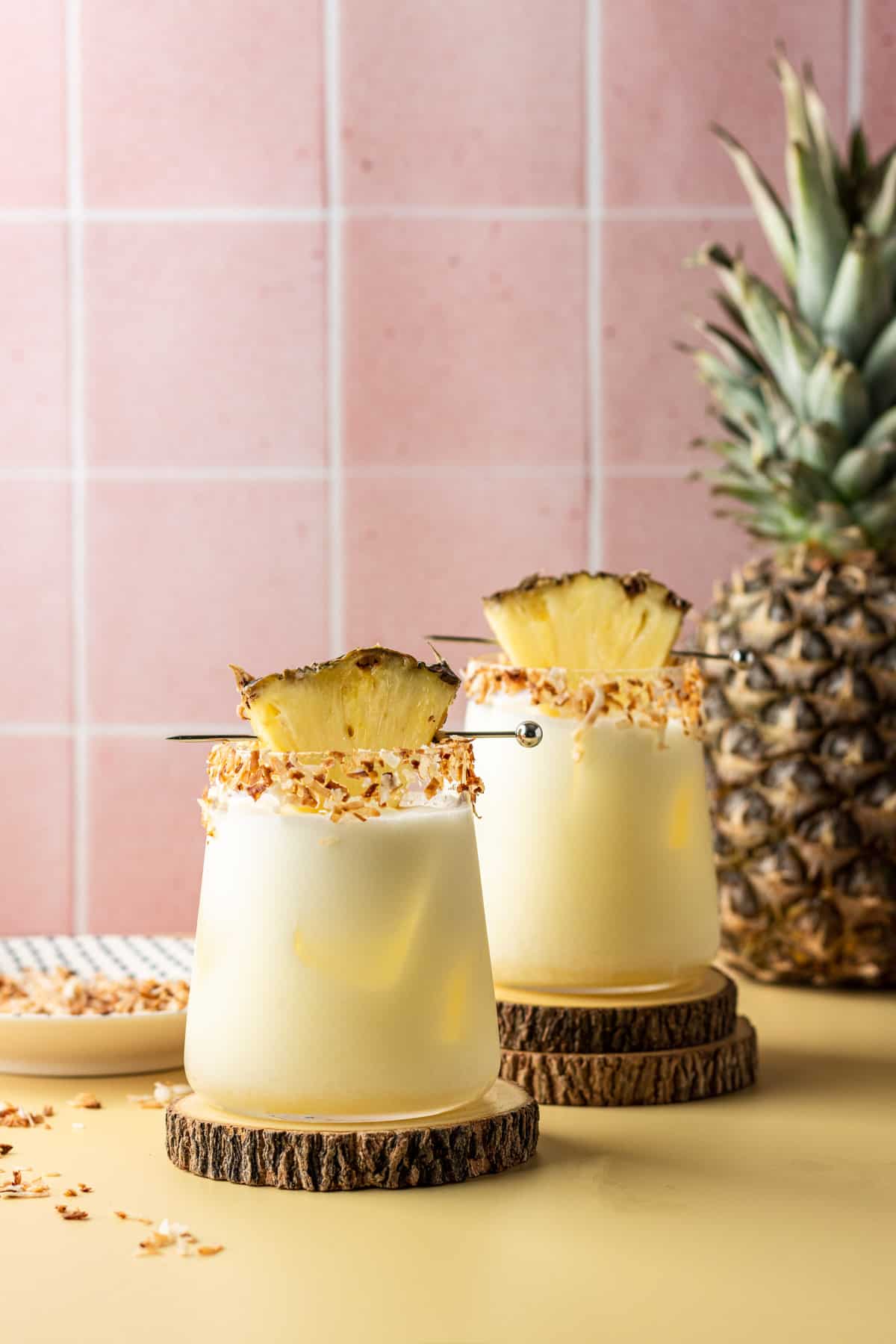 Two pineapple coconut margaritas in glasses garnished with toasted coconut and a pineapple wedge on wooden coasters.