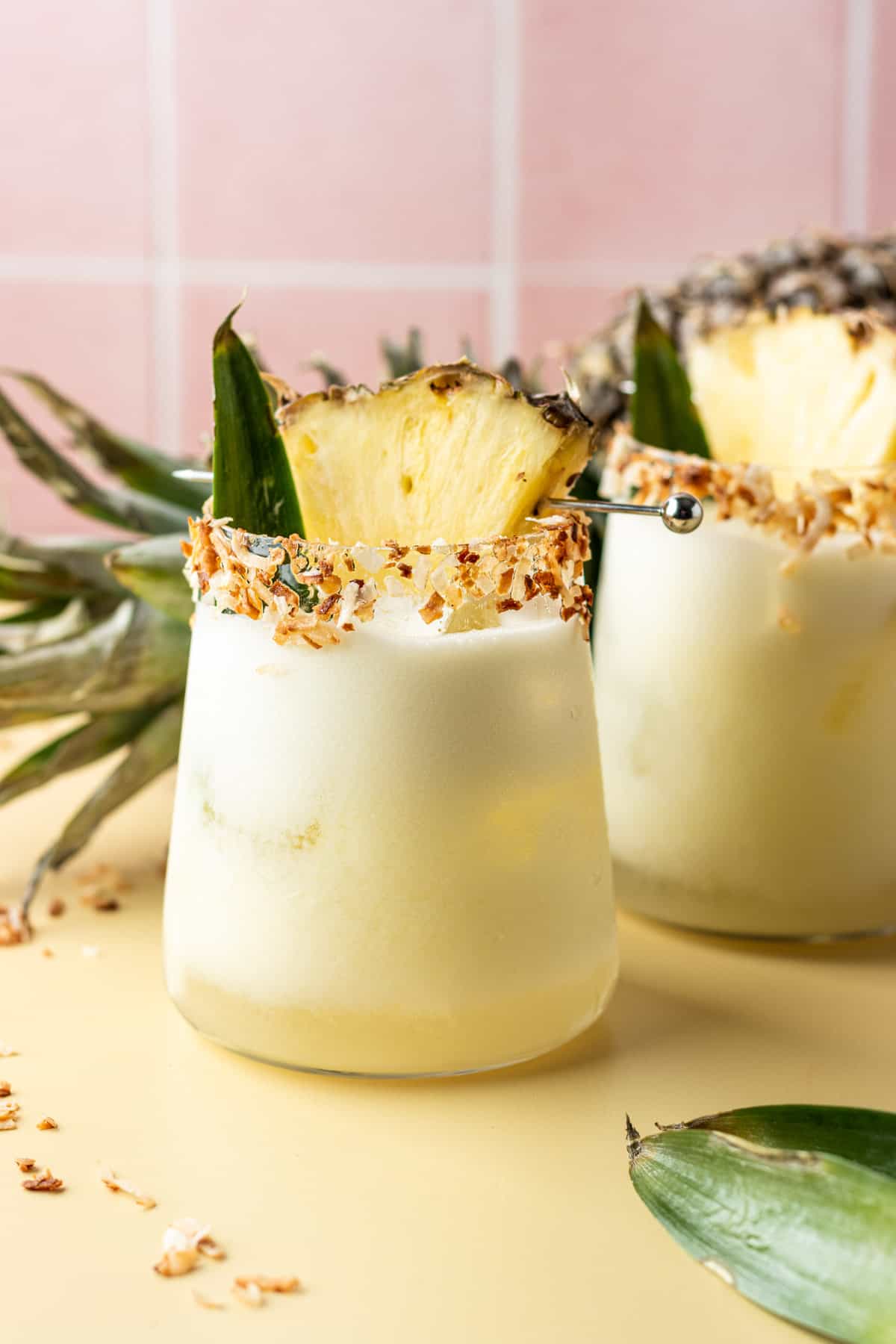 Two pineapple coconut margaritas in glasses garnished with toasted coconut flakes and a pineapple wedge.