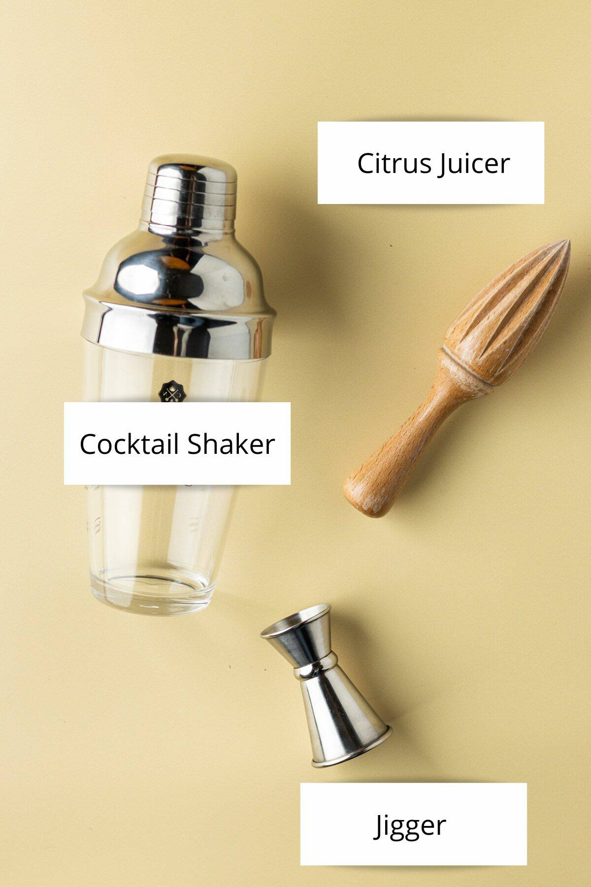 Overhead view of a cocktail shaker, citrus juicer and jigger on a yellow backdrop.