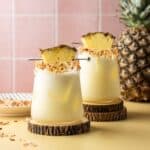 Two pineapple coconut margaritas in glasses on wooden coasters garnished with toasted coconut and a pineapple wedge.