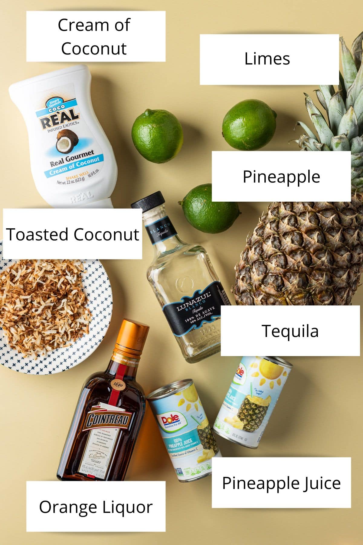 Overhead view of a bottle of cream of coconut, limes, pineapple, toasted coconut, tequila, cointreau and pineapple juice.