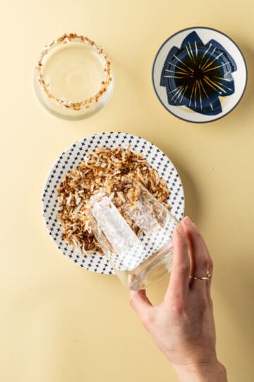 Rim of a glass being dipped into a plate of toasted coconut flakes.