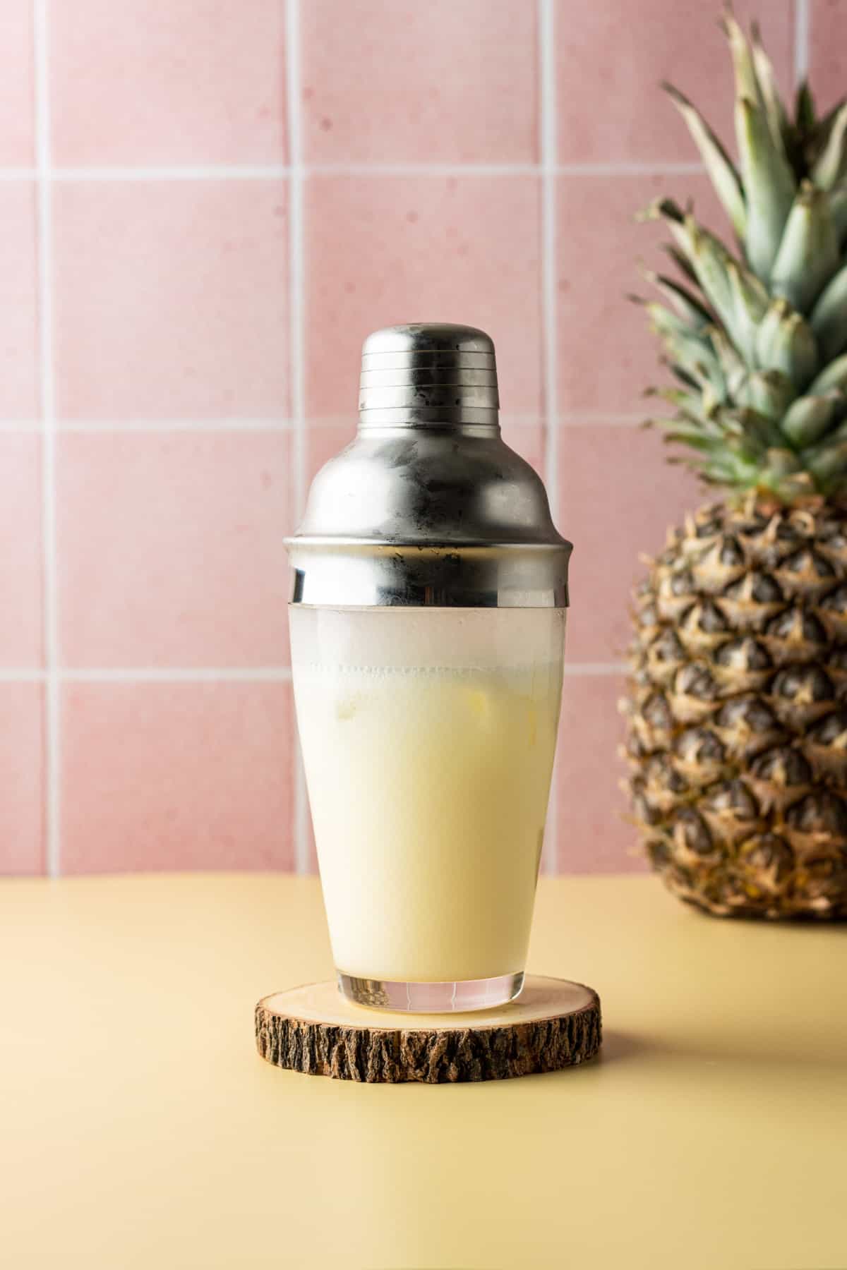 Coconut pineapple margarita in a cocktail shaker after being shaken standing on a wooden coaster.