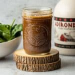Maple balsamic dressing in a jar on two wooden coasters with a jug of maple syrup and bowl of spinach.