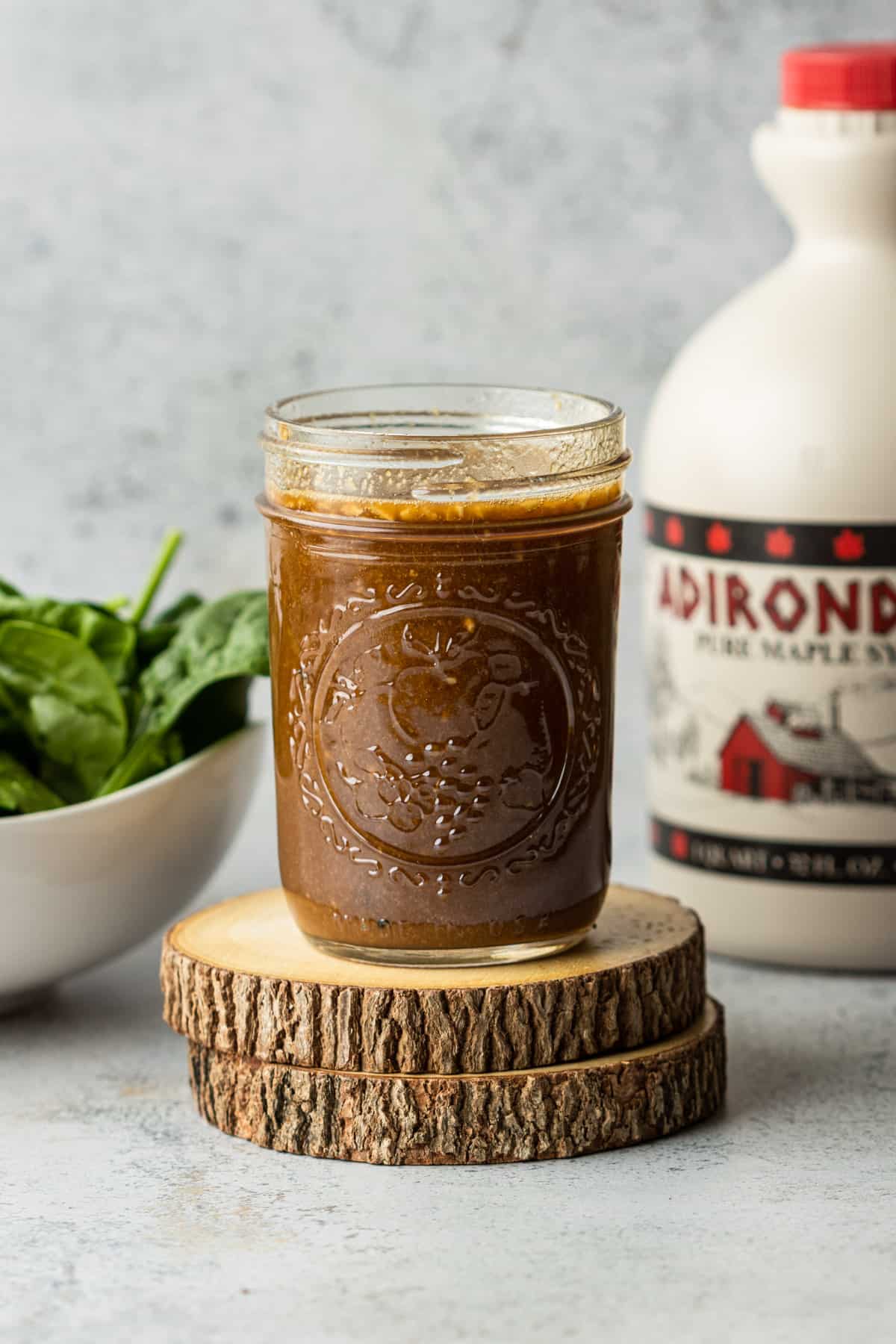 Sweet maple balsamic dressing in a jar on two wooden coasters with a jug of maple syrup and bowl of spinach.
