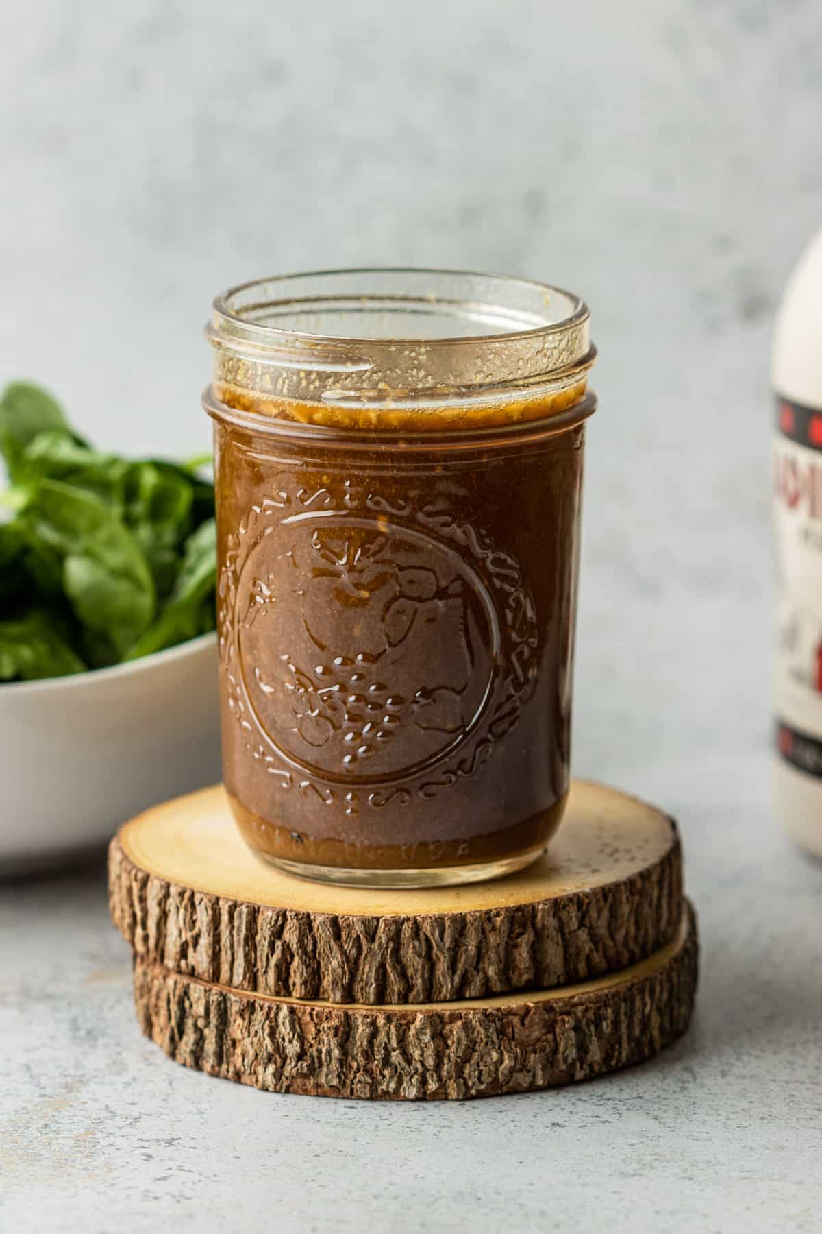 Homemade balsamic dressing in a jar on two wooden coasters with a jug of maple syrup and bowl of spinach.