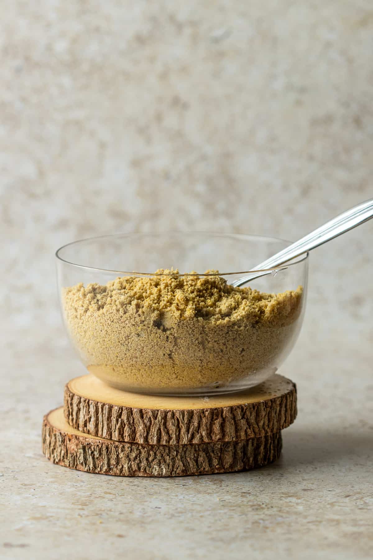 Homemade vegan parmesan cheese in a clear bowl with a spoon on two wooden coasters.