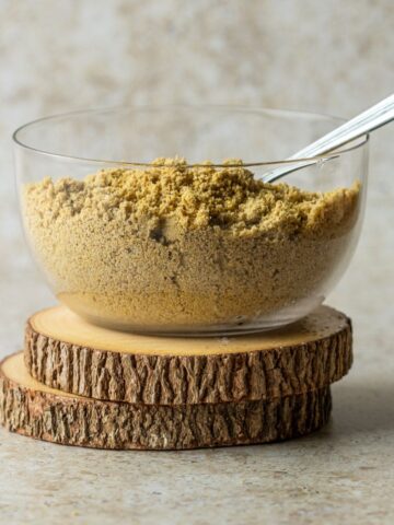 Homemade vegan parmesan cheese in a clear bowl with a spoon on two wooden coasters.