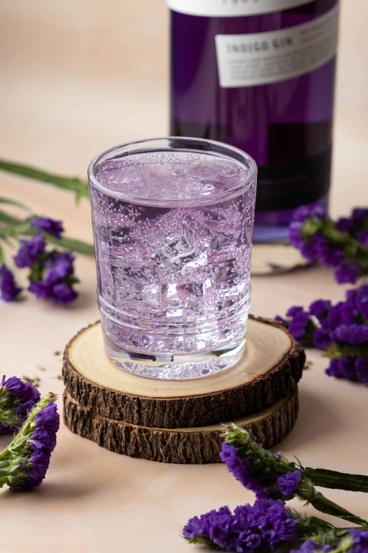 Lavender gin and tonic on two wooden coasters with Empress gin bottle in the background and purple flowers.