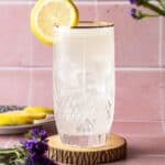 Tall glass of lavender vodka lemonade garnished with a lemon wheel on a wooden coaster with purple flowers around.