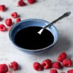 Raspberries scattered around a small blue bowl with a spoon filled with raspberry balsamic sauce.