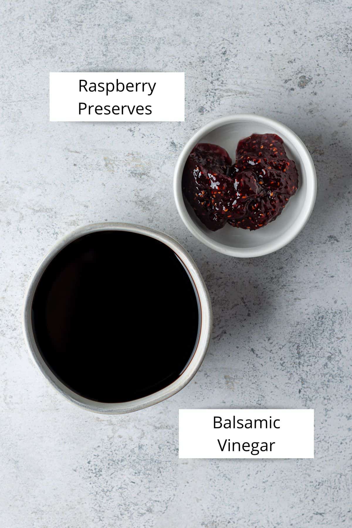 A small white bowl filled with raspberry preserves and a small grey bowl filled with balsamic vinegar.
