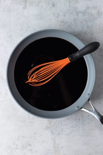 A frying pan with balsamic vinegar and raspberry preserves and an orange colored whisk.