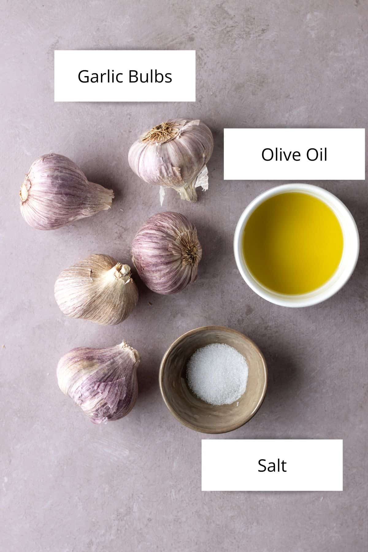 Five whole garlic bulbs, a white bowl of olive oil and a tan bowl of salt.