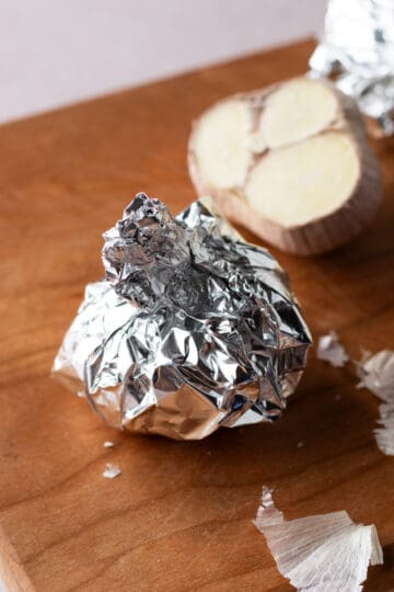 A garlic head wrapped up in foil on a wooden cutting board.