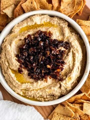 A close up of humus in a white bowl with caramelized onions of top and pita chips scattered around the bowl.