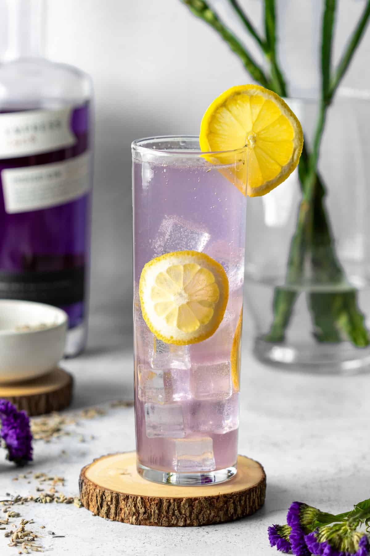 A tall glass on a wooden coaster with a purple collins drink garnished with lemon wheels and flowers.