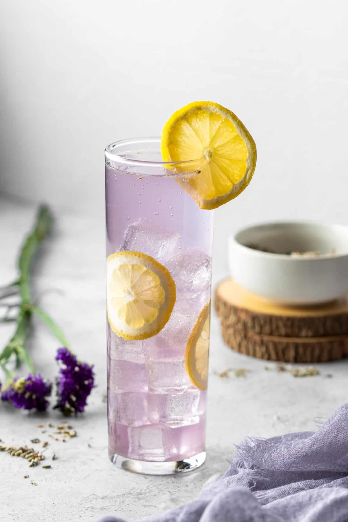 A Collins glass with a purple gin collins garnished with lemon wheels with purple flowers and a bowl of lavender.